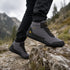 A person wearing dark gray and black hiking boots with yellow accents, standing on a rocky mountain path with a blurred natural background.