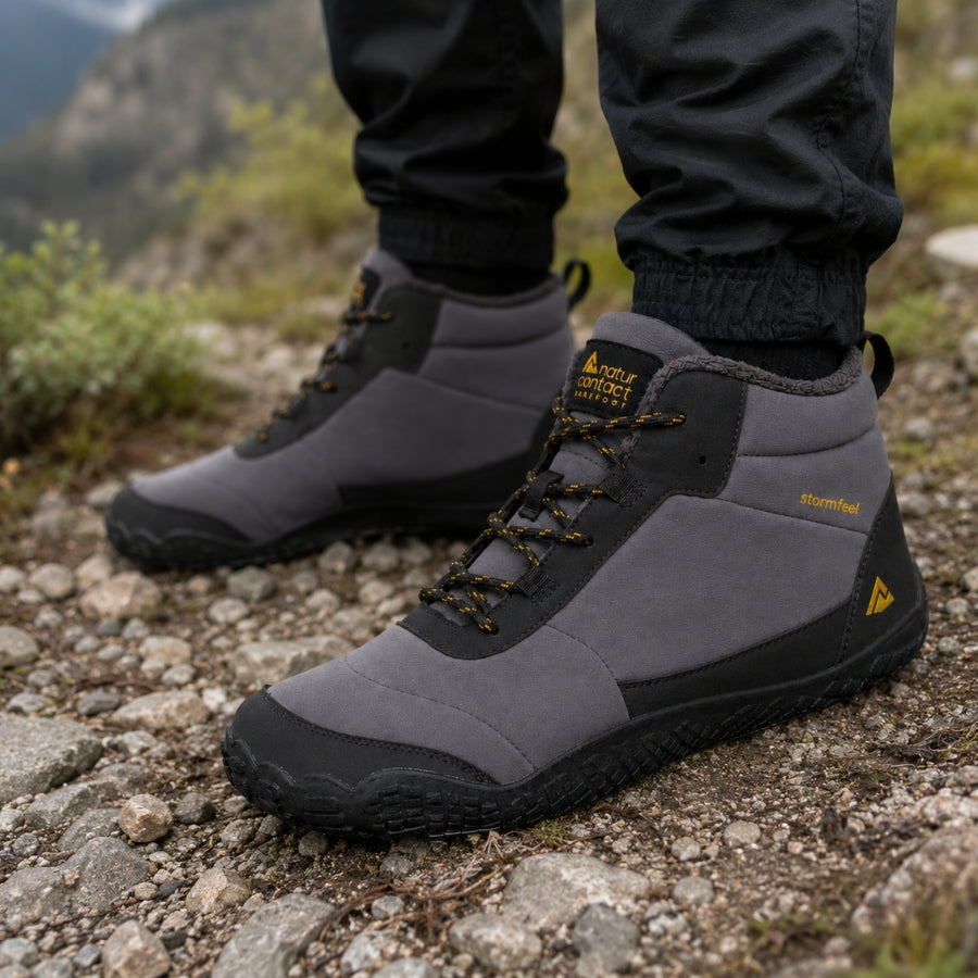 A pair of gray and black mid-cut hiking boots with yellow accents, shown on a rocky outdoor trail with a person's legs in black pants visible above the boots.