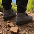 Rear view of grey Naturcontact outdoor boots on a rocky dirt path, showing the brand’s triangular heel logo and padded ankle design