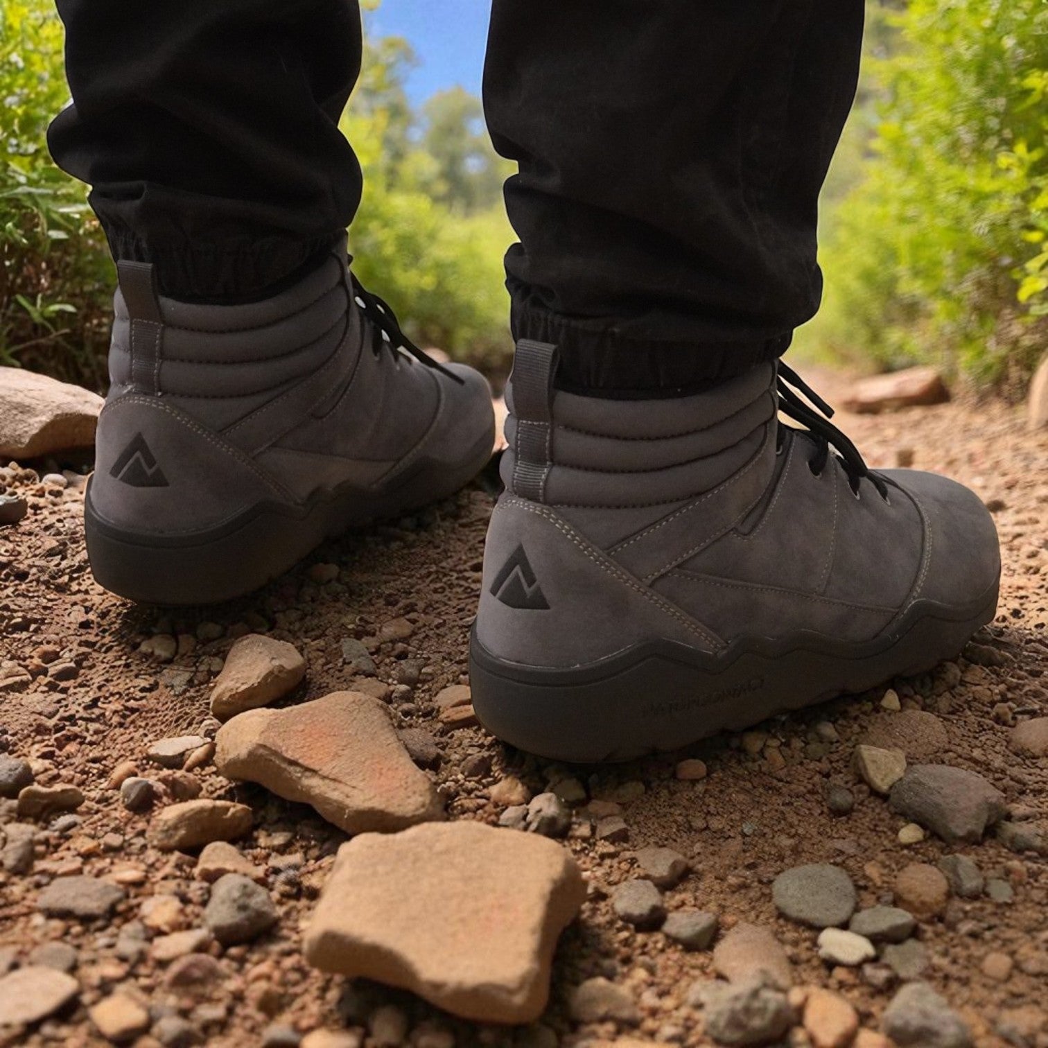 Rear view of grey Naturcontact outdoor boots on a rocky dirt path, showing the brand’s triangular heel logo and padded ankle design