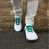 Front view of white leather barefoot sneakers with vibrant green laces and gum soles, worn with beige trousers against a stone wall background.