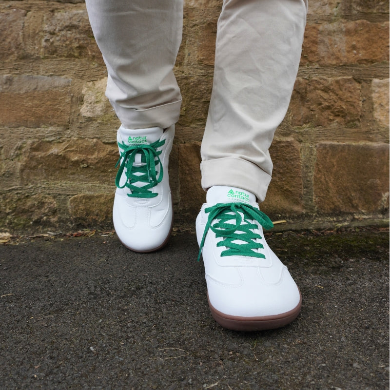 Front view of white leather barefoot sneakers with vibrant green laces and gum soles, worn with beige trousers against a stone wall background.