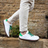Side profile of a person walking on a stone ledge wearing white leather barefoot sneakers with green laces, green heel tabs, and gum soles.