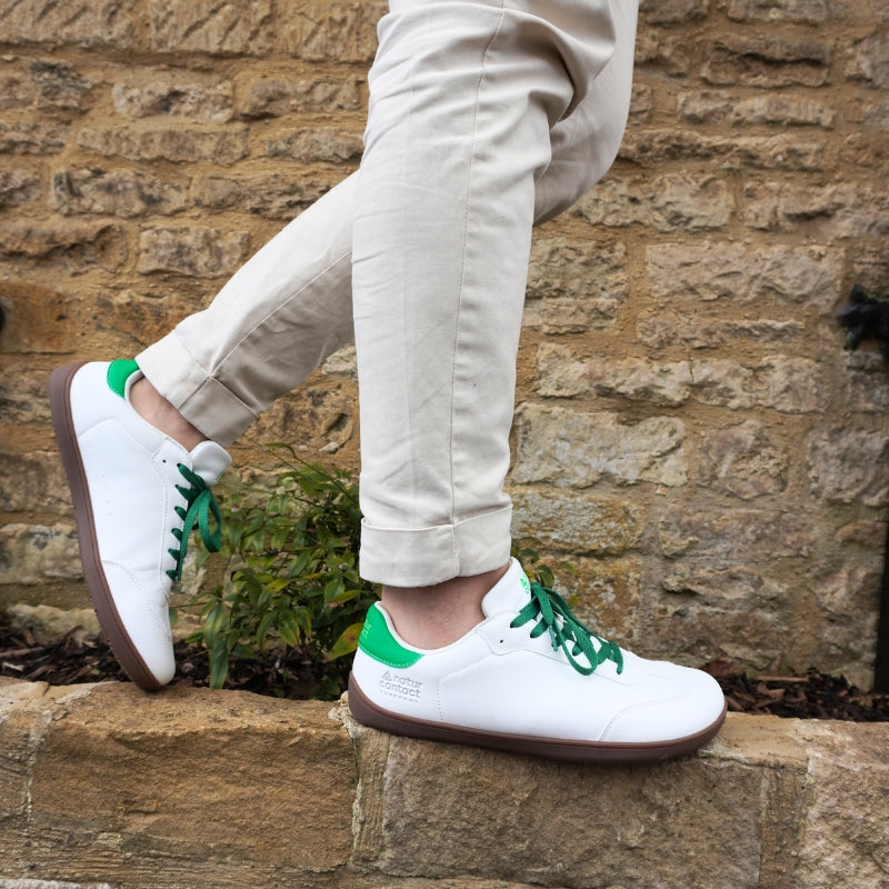 Side profile of a person walking on a stone ledge wearing white leather barefoot sneakers with green laces, green heel tabs, and gum soles.