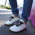 Side view of a person wearing white leather barefoot sneakers with black laces and gum soles, paired with blue jeans in an outdoor urban setting.