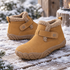 A pair of tan winter ankle boots with fluffy cream lining and hook-and-loop straps, placed on a snowy wooden deck outdoors.