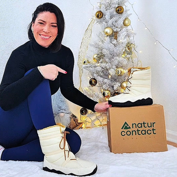 Woman wearing white winter boots pointing at a 'natur contact' box with a decorated Christmas tree in the background.